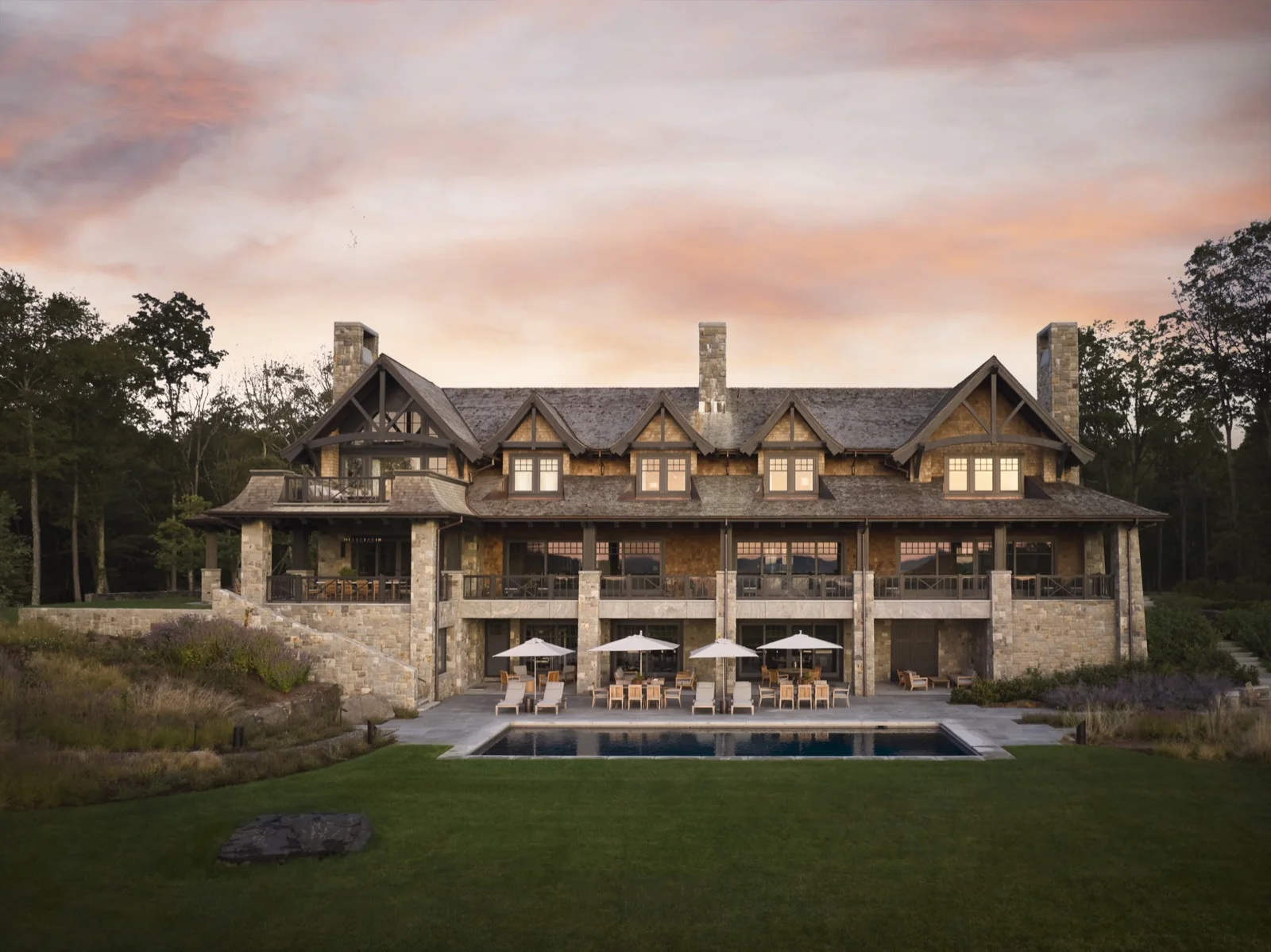 Stone and shingle mountain residence at dusk with a terraced lawn and pool, built by Hobbs, Inc.