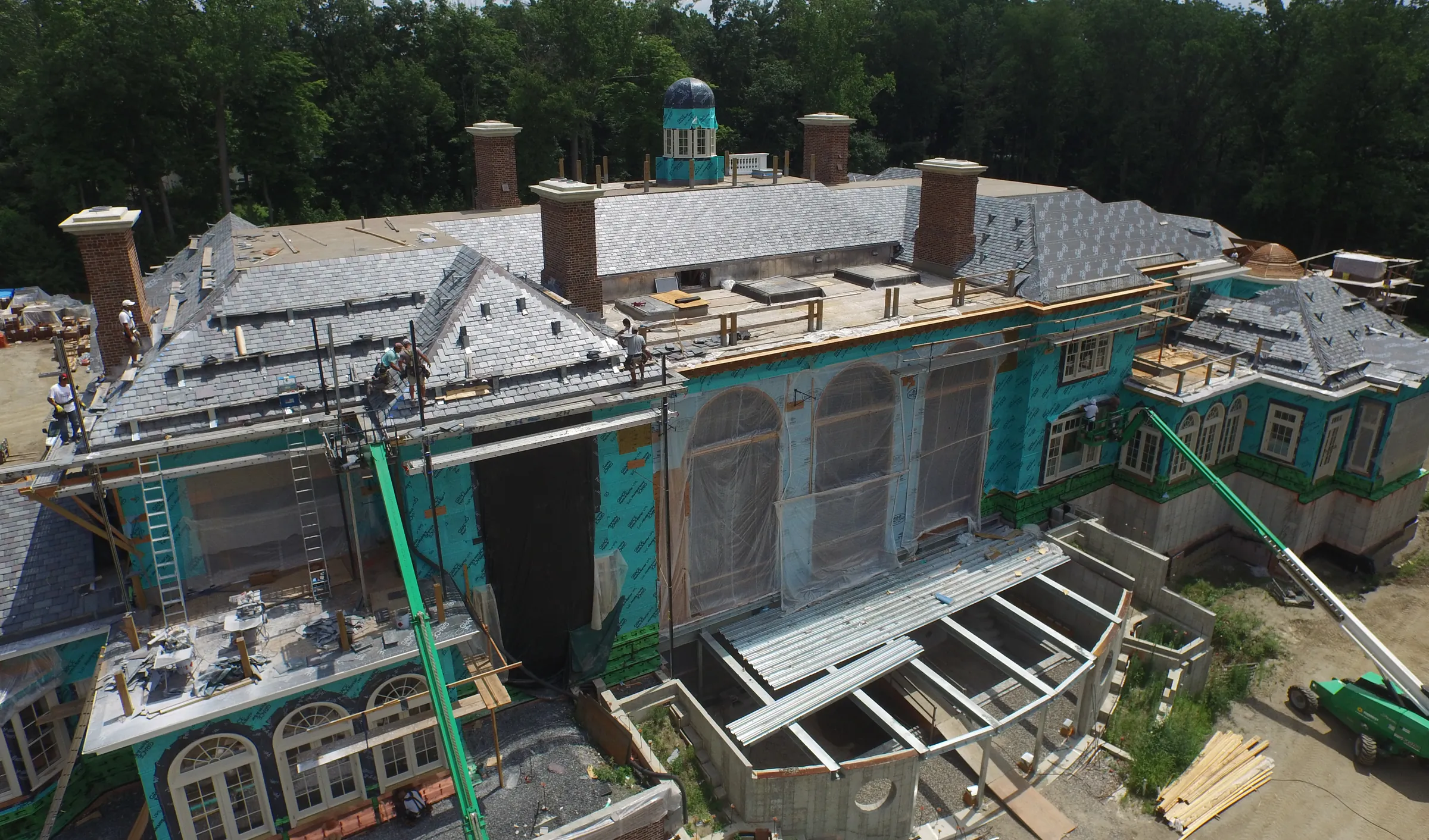Slate roofing, brick chimneys and a copper cupola going on over a wrapped facade on a Hobbs custom home, seen from above